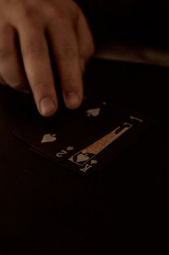 A hand is touching a stack of playing cards laid out on a dark surface. The cards are primarily black, featuring heart suits, including the king and jack. The scene is dimly lit, giving it an intimate or mysterious atmosphere.