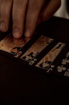 A hand gently resting on a spread of playing cards, featuring black suits and gold, ornate designs. The setting appears to be dimly lit, giving off a sophisticated and mysterious atmosphere.
