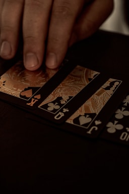 A hand gently resting on a spread of playing cards, featuring black suits and gold, ornate designs. The setting appears to be dimly lit, giving off a sophisticated and mysterious atmosphere.