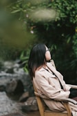 A person wearing a beige coat and sunglasses sits on a wooden chair, gazing towards a lush, green natural backdrop with a stream flowing nearby.