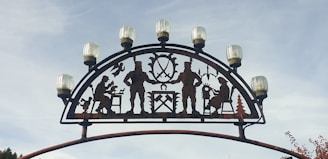 An arch with metal silhouettes depicting people engaged in various activities such as playing an instrument and woodworking is topped with evenly spaced decorative lights. The backdrop is a clear sky.