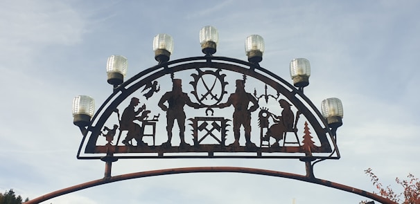 An arch with metal silhouettes depicting people engaged in various activities such as playing an instrument and woodworking is topped with evenly spaced decorative lights. The backdrop is a clear sky.