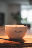 A steaming bowl of noodles placed on a wooden surface with chopsticks lying next to it. The bowl is white and has the word 'Noodles' printed on it. The background is softly blurred, giving a warm and cozy atmosphere.