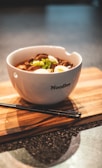 A bowl of noodles garnished with slices of boiled eggs, green onions, and what appears to be pieces of meat. The white bowl has the word 'Noodles' printed on it and is placed on a wooden board. Black chopsticks are also resting on the board beside the bowl.