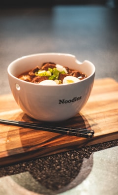 A bowl of noodles garnished with slices of boiled eggs, green onions, and what appears to be pieces of meat. The white bowl has the word 'Noodles' printed on it and is placed on a wooden board. Black chopsticks are also resting on the board beside the bowl.