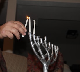 A hand is lighting candles on a menorah, which is a candelabrum typically used in Jewish rituals. The menorah is made of metal with tree branch-like holders. One candle has already been lit, and others are unlit.