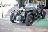 A classic vintage car is parked on a city street, showcasing its polished dark green exterior with a large front grille and distinctive round headlights. The car bears a European license plate and is positioned next to a storefront with clothing displayed in the window.