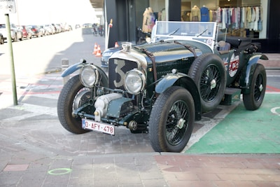 A classic vintage car is parked on a city street, showcasing its polished dark green exterior with a large front grille and distinctive round headlights. The car bears a European license plate and is positioned next to a storefront with clothing displayed in the window.