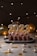 A festive cake adorned with chocolate decorations and candles shaped as numbers spelling out 2023 sits on a plate. Warm string lights and balloons in shades of gold and silver create a celebratory atmosphere. Scattered golden confetti and small shiny decorations are spread across the surface.