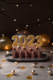 A festive cake adorned with chocolate decorations and candles shaped as numbers spelling out 2023 sits on a plate. Warm string lights and balloons in shades of gold and silver create a celebratory atmosphere. Scattered golden confetti and small shiny decorations are spread across the surface.