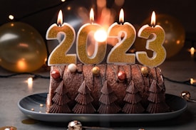 A decorated chocolate cake with chocolate tree-shaped designs sits on a plate. Large gold candles spelling out '2023' are lit on top of the cake. Surrounding the cake are festive elements like balloons and string lights, creating a celebratory atmosphere.