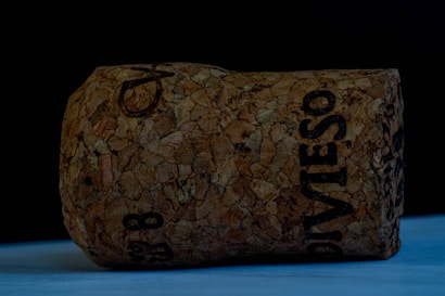 A large champagne cork with intricate textures and visible imprinted text lying on a smooth surface. The background is a dark, solid color, creating contrast with the lighter cork material.