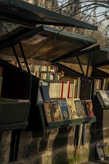 Several open bookshelves are filled with various books, located outdoors under a canopy of trees. The books are neatly arranged and include both large and small volumes, with some featuring illustrations on their covers. Below the shelves, a couple of umbrellas are stored upright against the stone wall.