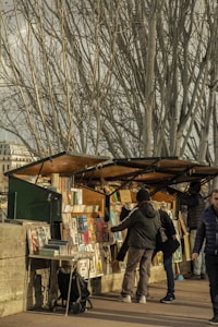 A street market bookstall with several open display boxes filled with books and prints is set against a backdrop of leafless trees. A few people are browsing through the items, dressed in winter clothing.
