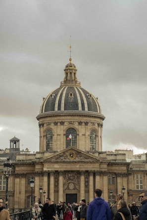 A grand architectural building with a large dome and a French flag visible. The facade includes ornate details and columns. In the foreground, a group of people are walking, indicating a popular or tourist area. The atmosphere is overcast with grey clouds in the sky.