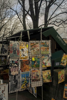 A street vendor stall displays a variety of vintage posters, including illustrations from Mickey Mouse, Tour de France, and other retro-style prints. The stall is set against a backdrop of leafless trees and a cloudy sky. The posters are arranged on a metal grid, showcasing diverse colors and themes.