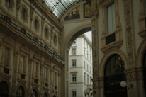 An ornate architectural interior featuring grand arches and intricate decorative details with classical paintings. The space is illuminated by natural light filtering through a glass ceiling. A luxury store is visible on the right side.