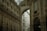 An ornate architectural interior featuring grand arches and intricate decorative details with classical paintings. The space is illuminated by natural light filtering through a glass ceiling. A luxury store is visible on the right side.