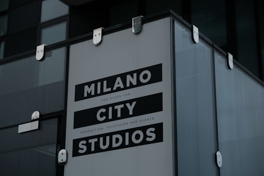 A large sign with bold text reads 'Milano City Studios'. The sign is attached to a modern building with glass panels and metallic fasteners. It appears to be part of a production or event location.
