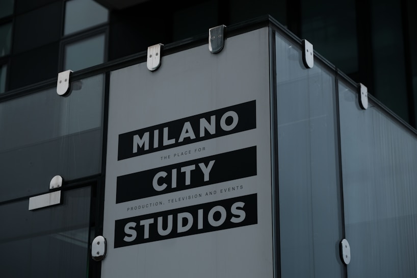 A large sign with bold text reads 'Milano City Studios'. The sign is attached to a modern building with glass panels and metallic fasteners. It appears to be part of a production or event location.