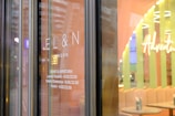 Glass door with the words 'EL&N London' displayed. The hours of operation are listed beneath, indicating Monday to Friday from 8.00 to 22.00, and Saturday, Sunday, and holidays from 9.00 to 22.00. The interior is visible with pastel colors and the word 'Adventure' prominently displayed.