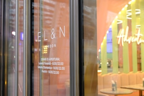 Glass door with the words 'EL&N London' displayed. The hours of operation are listed beneath, indicating Monday to Friday from 8.00 to 22.00, and Saturday, Sunday, and holidays from 9.00 to 22.00. The interior is visible with pastel colors and the word 'Adventure' prominently displayed.