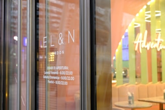Glass door with the words 'EL&N London' displayed. The hours of operation are listed beneath, indicating Monday to Friday from 8.00 to 22.00, and Saturday, Sunday, and holidays from 9.00 to 22.00. The interior is visible with pastel colors and the word 'Adventure' prominently displayed.