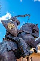 A close-up view of a metallic statue of a knight on horseback. The knight holds a shield bearing a gold emblem, and a flag above the knight carries the letters 'UCF'. The background shows a clear blue sky with white clouds and part of a building.