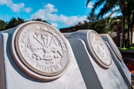 Several large stone plaques engraved with military emblems and text are aligned outdoors in a landscaped area. The surrounding area features green palm trees and other greenery under a bright blue sky with scattered clouds.