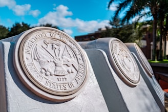 Several large stone plaques engraved with military emblems and text are aligned outdoors in a landscaped area. The surrounding area features green palm trees and other greenery under a bright blue sky with scattered clouds.