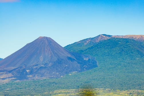A volcanic mountain with a steep, dark ash-covered slope rises prominently next to a lush green, tree-covered hill. The sky is clear and blue, creating a vivid contrast with the earthy tones of the volcano and the greenery.