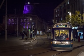 A nighttime urban scene features a tram labeled 'Phoenix Park' traveling down the street. The well-lit area includes a historic building with purple lighting and a clock on the dome. Pedestrians are visible on the sidewalk, with street lamps illuminating the scene and a few storefronts.
