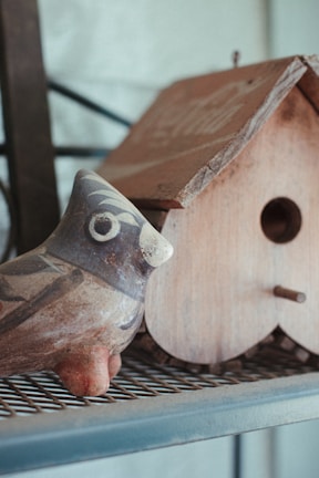 A rustic ceramic ornament shaped like a bird, glazed in earthy tones and perched on a wooden shelf.