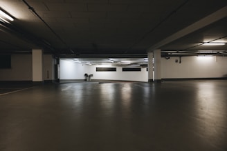 A dimly lit underground parking garage with concrete floors and walls. The space is expansive and mostly empty, illuminated by fluorescent lights. Piping and ventilation systems are visible on the ceiling.