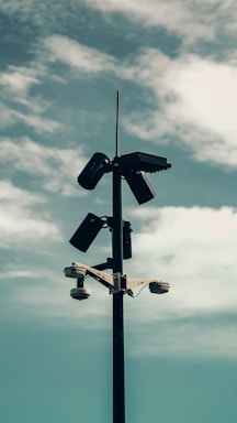 A tall pole with multiple surveillance cameras and lights mounted at the top under a cloudy sky. The angle creates an imposing and somewhat mysterious atmosphere.