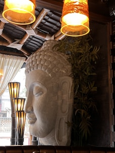 A large stone sculpture of a serene face occupies a prominent place in a warmly lit indoor setting. The background features bamboo structures and leafy plants, contributing to an earthy and tranquil atmosphere. Multiple light fixtures cast a soft glow, creating an inviting ambiance.