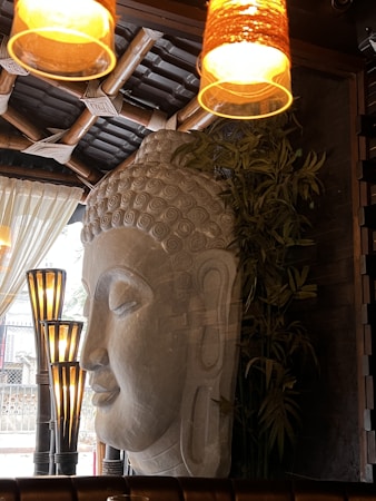 A large stone sculpture of a serene face occupies a prominent place in a warmly lit indoor setting. The background features bamboo structures and leafy plants, contributing to an earthy and tranquil atmosphere. Multiple light fixtures cast a soft glow, creating an inviting ambiance.