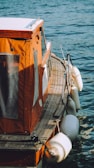 The image showcases a small boat with an orange canopy and wooden deck floating on a body of water. White fenders hang along the side, and the water is a rich blue color, gently rippling against the boat.