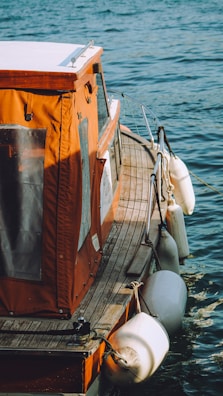 The image showcases a small boat with an orange canopy and wooden deck floating on a body of water. White fenders hang along the side, and the water is a rich blue color, gently rippling against the boat.