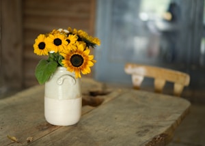 A rustic setting with a bouquet of vibrant sunflowers arranged in a ceramic vase on a wooden table. The background features a soft-focus interior with a wooden chair and muted colors.