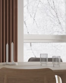 A minimalist interior features a light wooden table with two unique tall spiral candle holders and two ribbed glass cups. A large window in the background shows a snowy scene with bare tree branches, giving a serene and tranquil feel. Brown vertical blinds partially cover one side of the window.