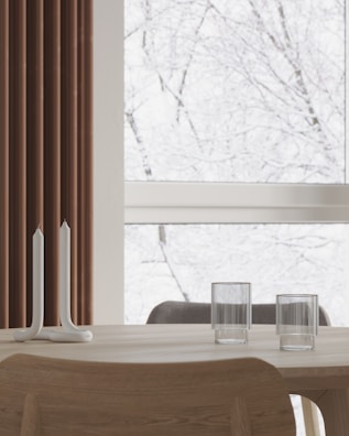 A minimalist interior features a light wooden table with two unique tall spiral candle holders and two ribbed glass cups. A large window in the background shows a snowy scene with bare tree branches, giving a serene and tranquil feel. Brown vertical blinds partially cover one side of the window.