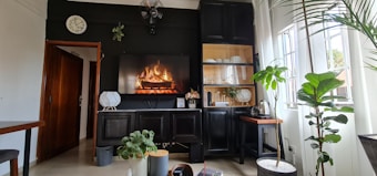 A cozy living room with a modern aesthetic features a digital fireplace displayed on a screen mounted on a dark wall. Surrounding it are dark wooden cabinets with glass doors showcasing dishes and glassware. There