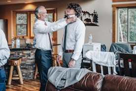 An older man is adjusting a younger man's tie in a cozy living room setting. The room features wooden furniture and decor, with large windows and a stone facade kitchen bar. Clothes and bottled water are visible around the room, giving a casual, preparatory vibe.