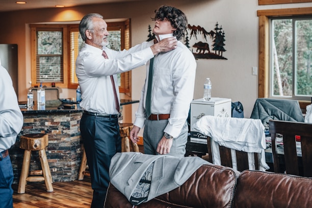 An older man is adjusting a younger man's tie in a cozy living room setting. The room features wooden furniture and decor, with large windows and a stone facade kitchen bar. Clothes and bottled water are visible around the room, giving a casual, preparatory vibe.
