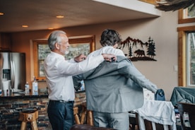 A man with gray hair is helping a younger man put on a suit jacket inside a cozy room with wooden furnishings. The room has a rustic feel with a stone countertop and a metal wall decoration of mountains and trees.