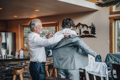 A man with gray hair is helping a younger man put on a suit jacket inside a cozy room with wooden furnishings. The room has a rustic feel with a stone countertop and a metal wall decoration of mountains and trees.