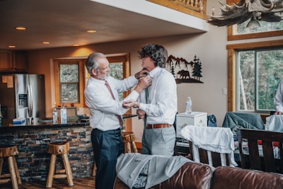 An older man is helping a younger man adjust his tie in a cozy, rustic kitchen with wooden furnishings and decor. There are two other individuals partially visible, one sitting on the left and another on the right. The area appears well-lit with natural light coming through the windows.
