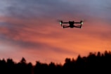 A drone is silhouetted against a vibrant orange and purple sunset sky, with faint green lights visible on its structure. The horizon shows a dark outline of trees or foliage adding to the contrast.