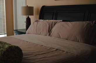 A neatly made bed with light brown sheets and pillows, situated next to a dark-colored side table with a decorative lamp. The room features soft lighting and neutral wall colors. A door and window with blinds are partially visible.